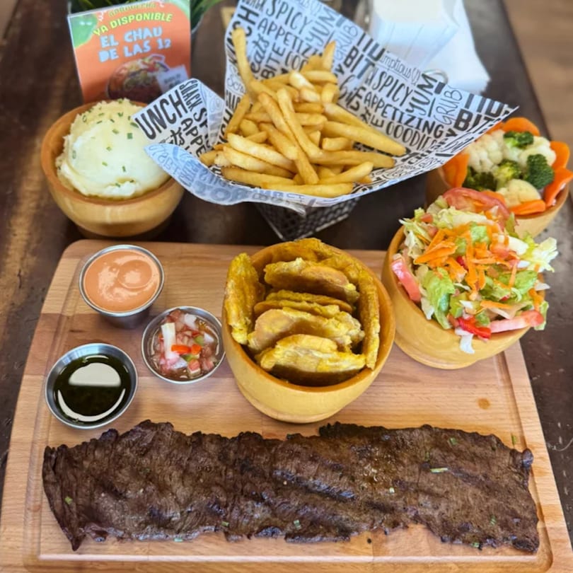 Grilled steak with fries, mashed potatoes, plantains, salad, and sauces on a wooden board.