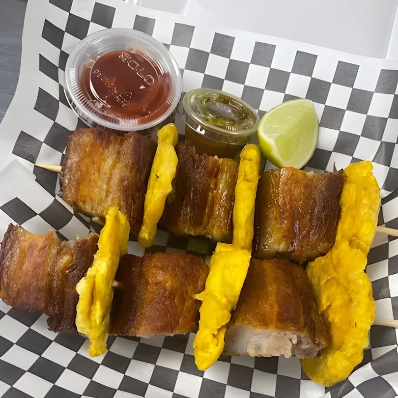 Two skewers of fried pork belly and plantains with dipping sauces and a lime wedge.