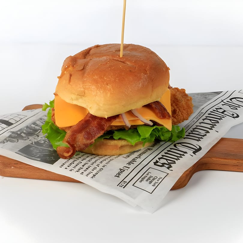 Grumpy G Restaurant's chicken burger with bacon, cheese, lettuce, and onions on newspaper-lined board.