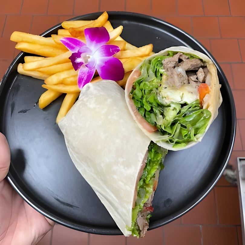 A black plate holds a steak and lettuce wrap with fries and a purple orchid garnish.