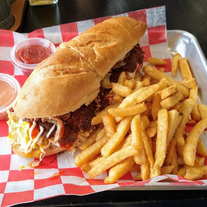 A Grumpy G Restaurant burger on a baguette with fries and dipping sauces on a metal tray.